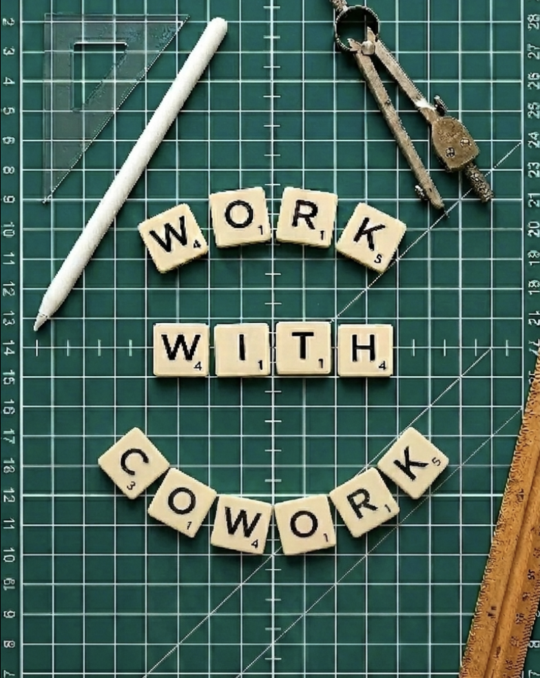 WORK WITH COWORK spelled out in scrabble tiles on a green cutting mat with rulers and a compass
